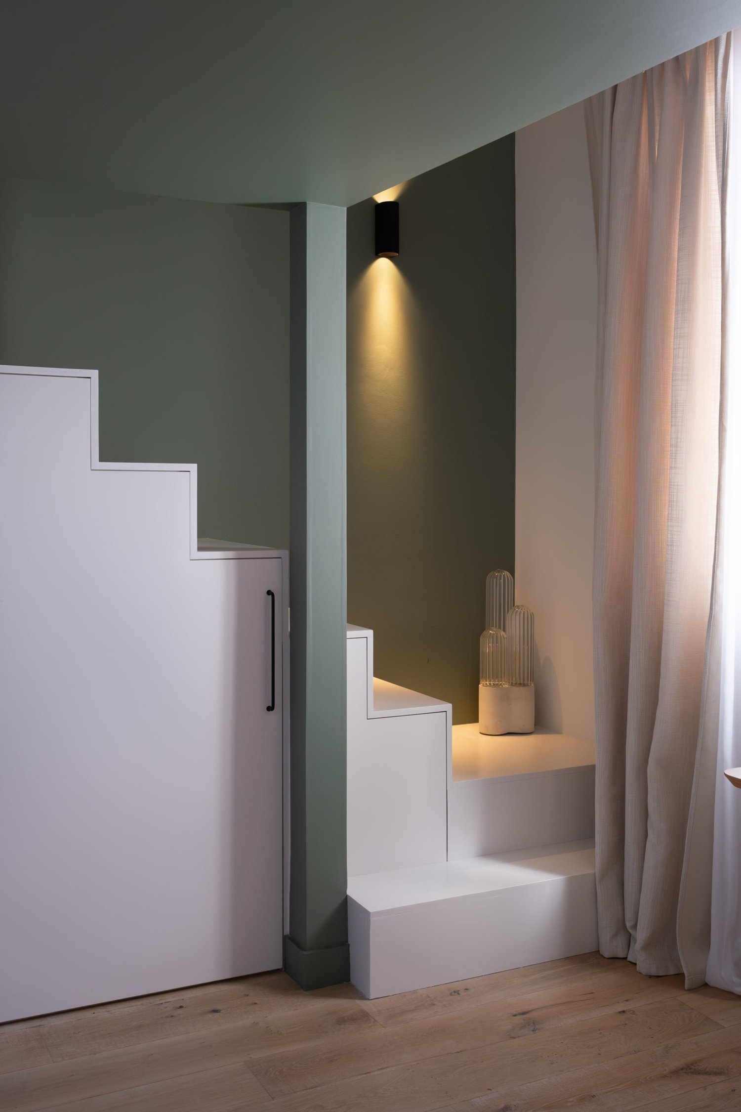 Placard intégré en escalier dans un intérieur moderne, avec un mur vert et une lumière encastrée. Vases décoratifs transparents en haut à droite, et une grande fenêtre avec rideaux en tissu clair.