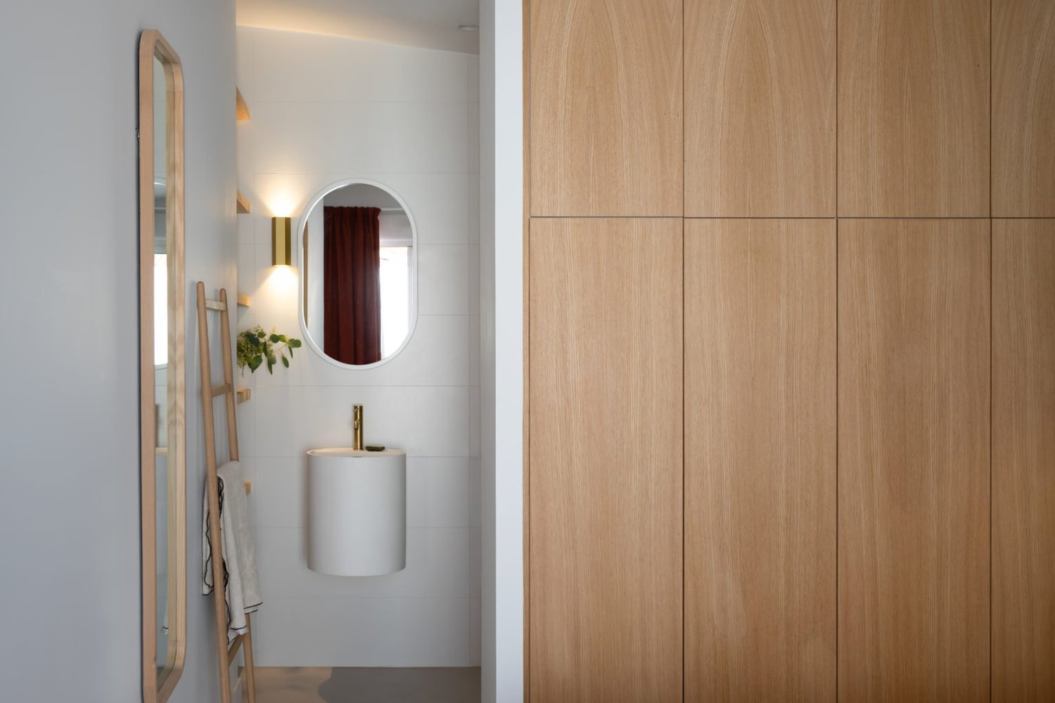 Salle de bain minimaliste avec un miroir ovale, un lavabo blanc avec robinet doré, une échelle en bois avec des serviettes, une plante verte, et un éclairage doux