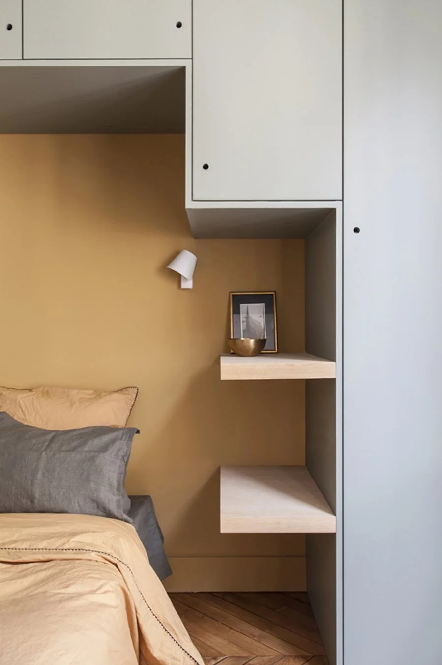 Un coin chambre avec un lit de couleur beige, deux coussins gris, un mur jaune, une lampe de lecture blanche, une étagère en bois avec un cadre photo, un bol doré, et un grand meuble gris avec des portes.