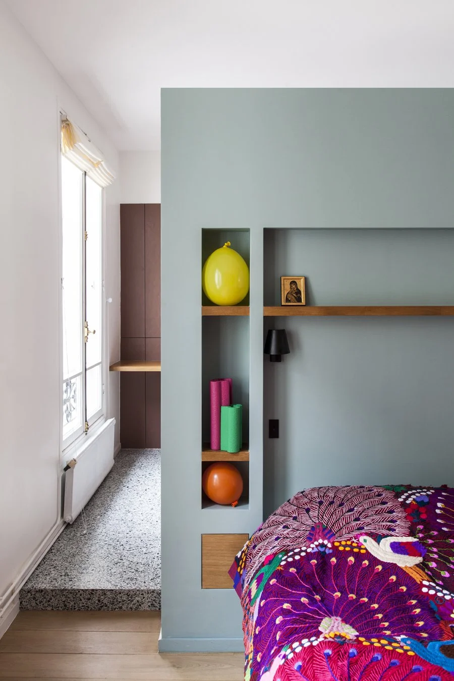 Une chambre avec un mur vert et des étagères en bois contenant des objets colorés, un lit avec une couverture multicolore, une fenêtre laissant entrer la lumière naturelle.