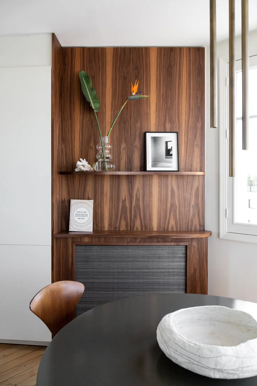 Un mur en bois avec une étagère, une grande carafe en verre contenant une feuille de plante tropicale et une fleur orange, une photo en noir et blanc, un livre, et une fenêtre à droite, avec une table ronde noire au premier plan et un bol blanc.