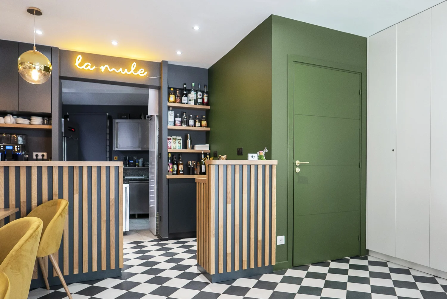 Intérieur d'un café ou restaurant avec une zone de bar en bois, un mur vert, un plafond blanc, et un sol en carreaux noir et blanc, avec un panneau lumineux 'la mule' en néon jaune.