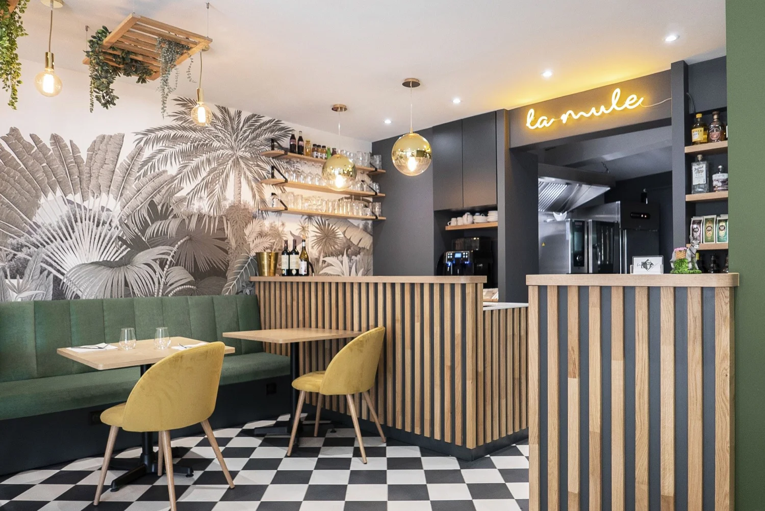 Intérieur d'un restaurant avec une décoration moderne. Mur avec une fresque de feuilles tropicales en noir et blanc, plafond avec des suspensions en forme de boulons en or, comptoir en bois avec rayures noires et en bois clair, fauteuils jaunes et ba