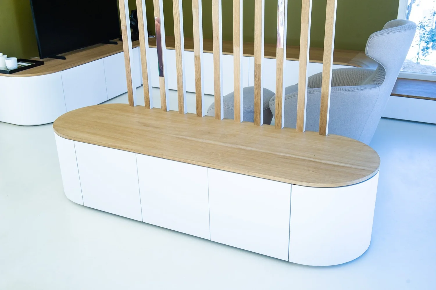 Banquette moderne avec dossier en bois et côtés blancs, située dans un salon lumineux avec fauteuil beige et télévision à écran plat