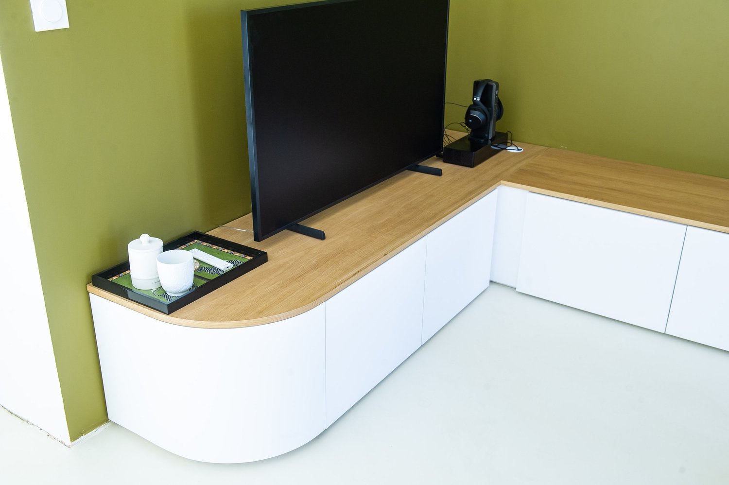 Espace de divertissement avec une télévision noire, un casque audio, un plateau avec une tasse blanche, un pot, un massicot, le tout sur un meuble en bois avec portes blanches, contre un mur vert