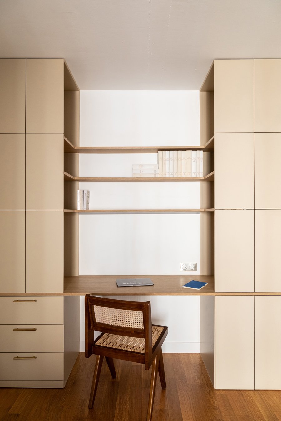 Bureau avec un bureau en bois, une chaise en bois avec cannage, une étagère encastrée blanche avec des livres, et une prise électrique sur le mur blanc.