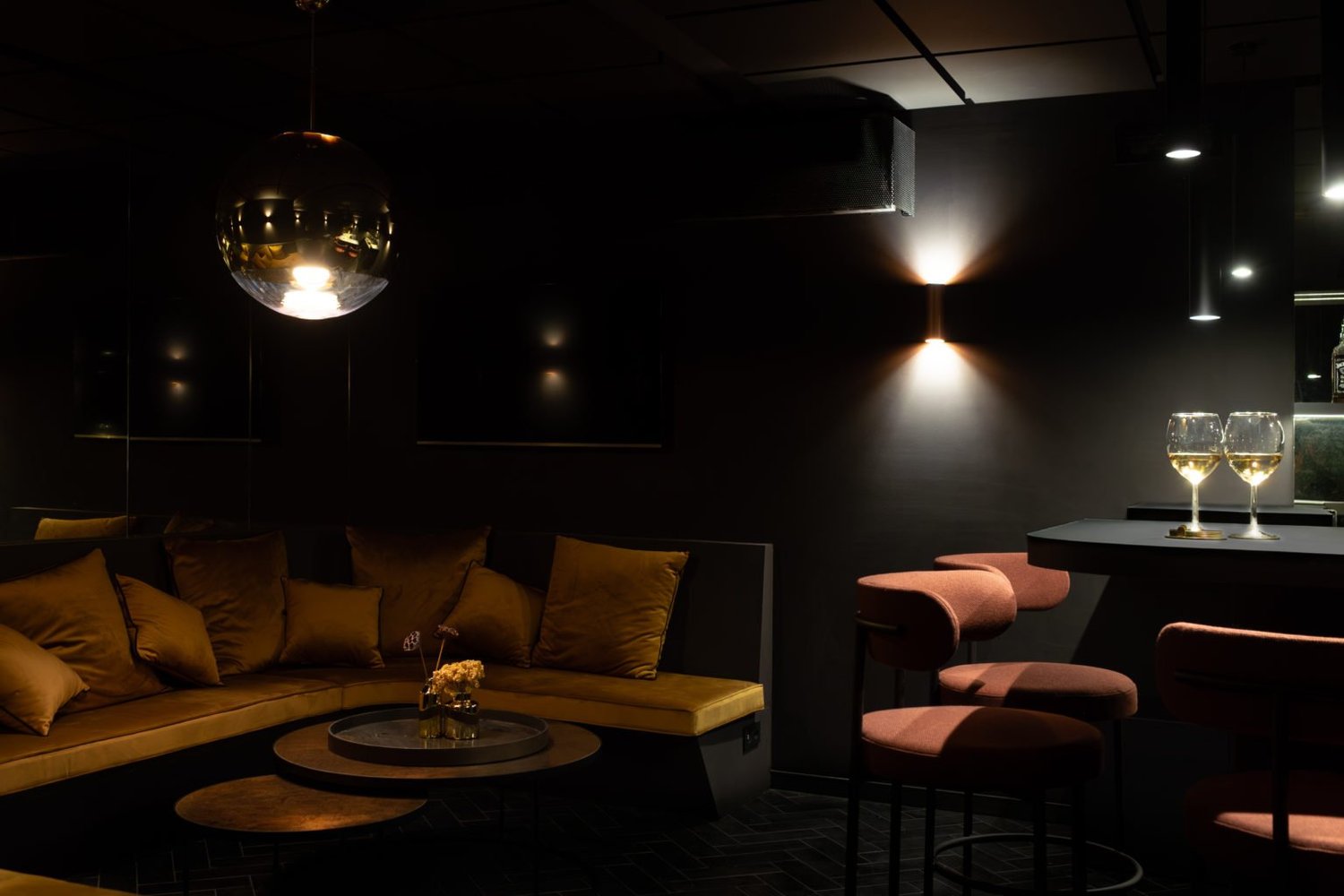 Intérieur d'un salon avec un canapé en velours jaune, des coussins, une table ronde avec une décoration florale, une table haute avec deux verres de vin blanc, un éclairage d'ambiance tamisé, et un miroir.