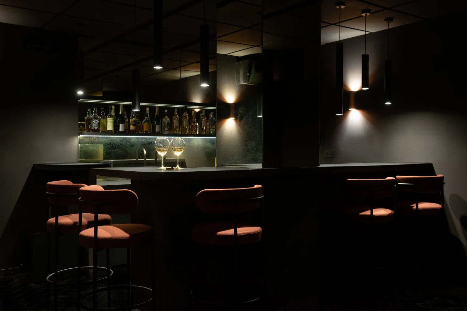 Un bar intérieur sombre avec des chaises rouges et deux verres de vin blanc sur le comptoir, des bouteilles d'alcool sur une étagère éclairée en arrière-plan.