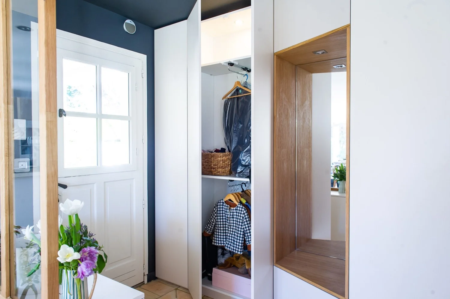 Un placard dans une entrée avec des vêtements suspendus, une caisse à linge, et des éléments décoratifs tels qu'un vase avec des fleurs.