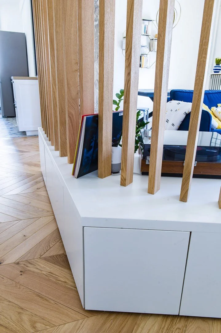 Vue intérieure d'un coin salon avec un divan bleu, des coussins, et des plantes. Séparation en bois avec livres posés.