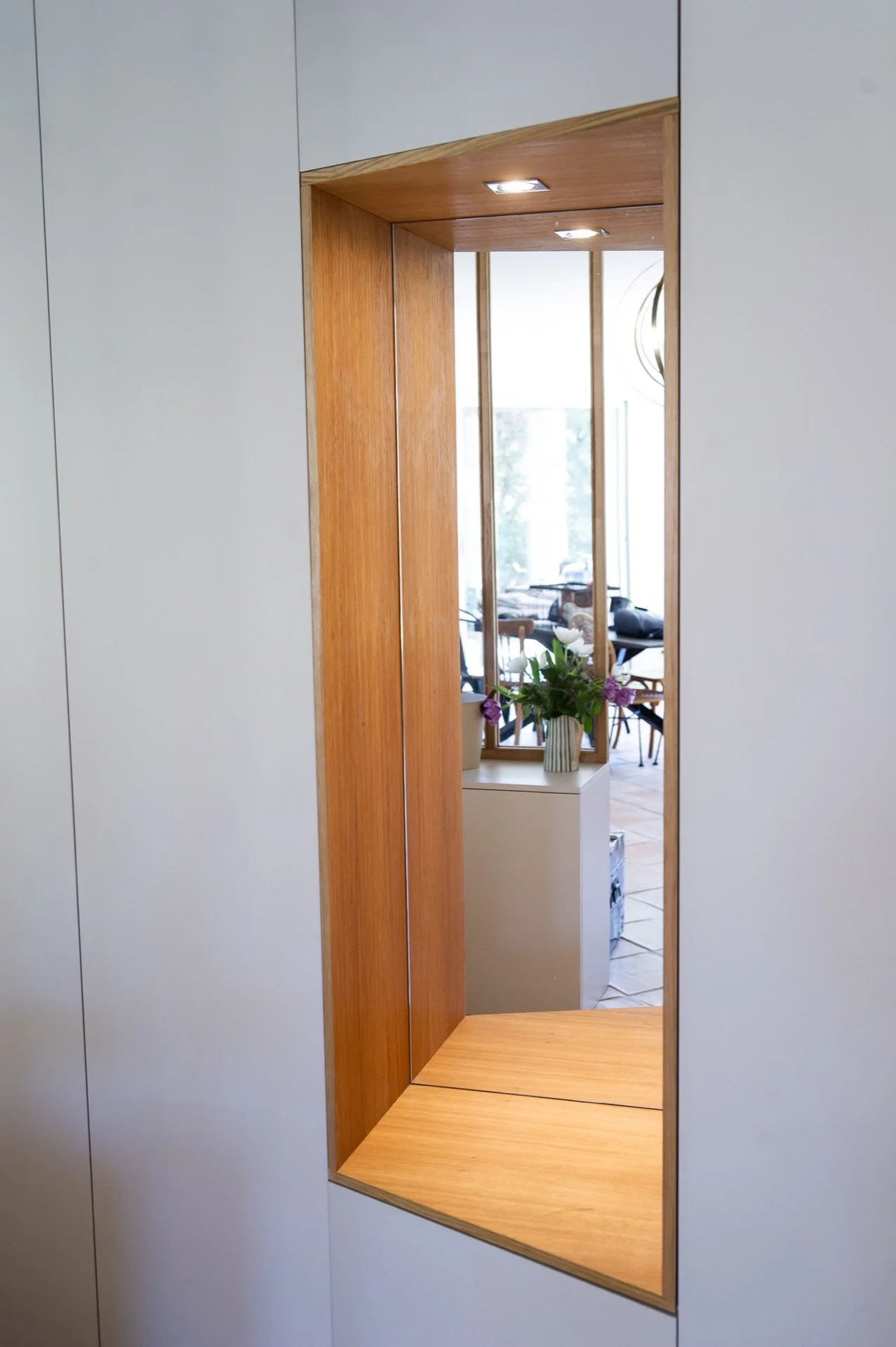 Un miroir encastré dans un mur blanc, avec un intérieur en bois visible, reflet d'une pièce avec des plantes et des meubles
