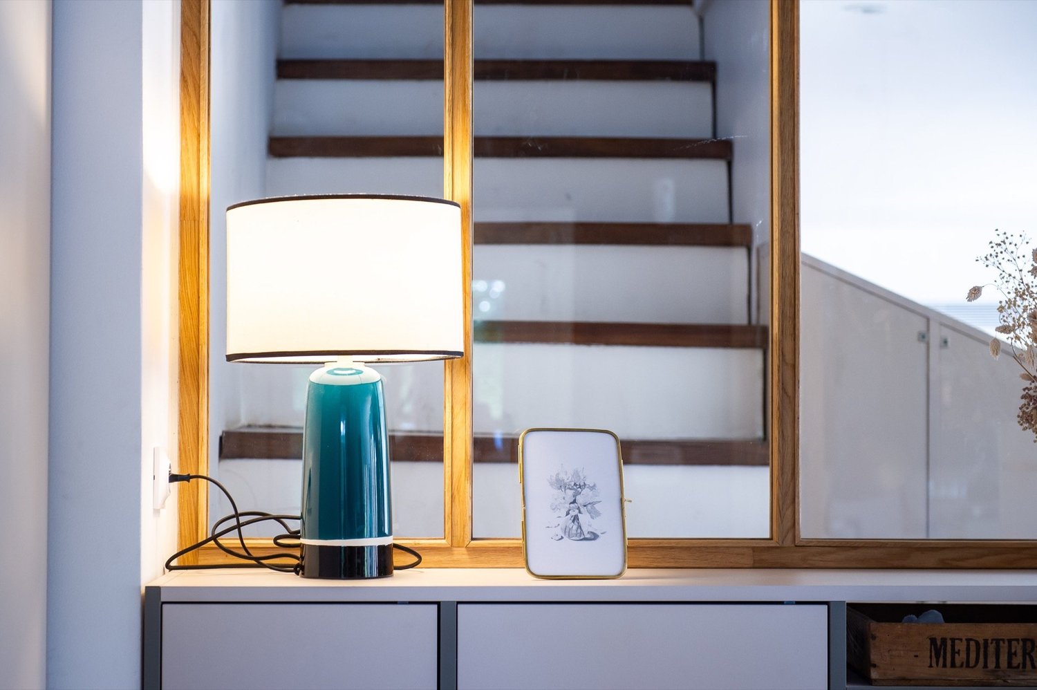 Lampe de table turquoise allumée, cadre en bois, petite image de décoration à côté, miroir derrière, meuble blanc en dessous, escalier en bois en arrière-plan, partie d'une pièce intérieure