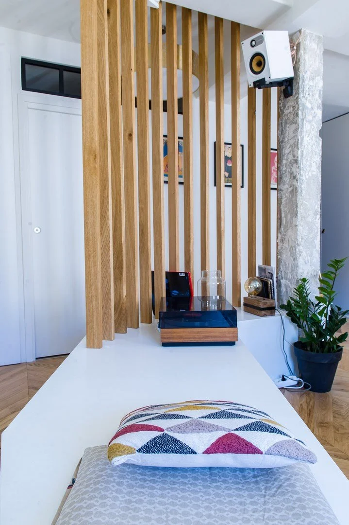 Vue d'un coin intérieur moderne avec un divan blanc, un coussin coloré, une plante en pot, un haut-parleur suspendu au plafond et une œuvre d'art sur le mur, séparés par un panneau en bois.