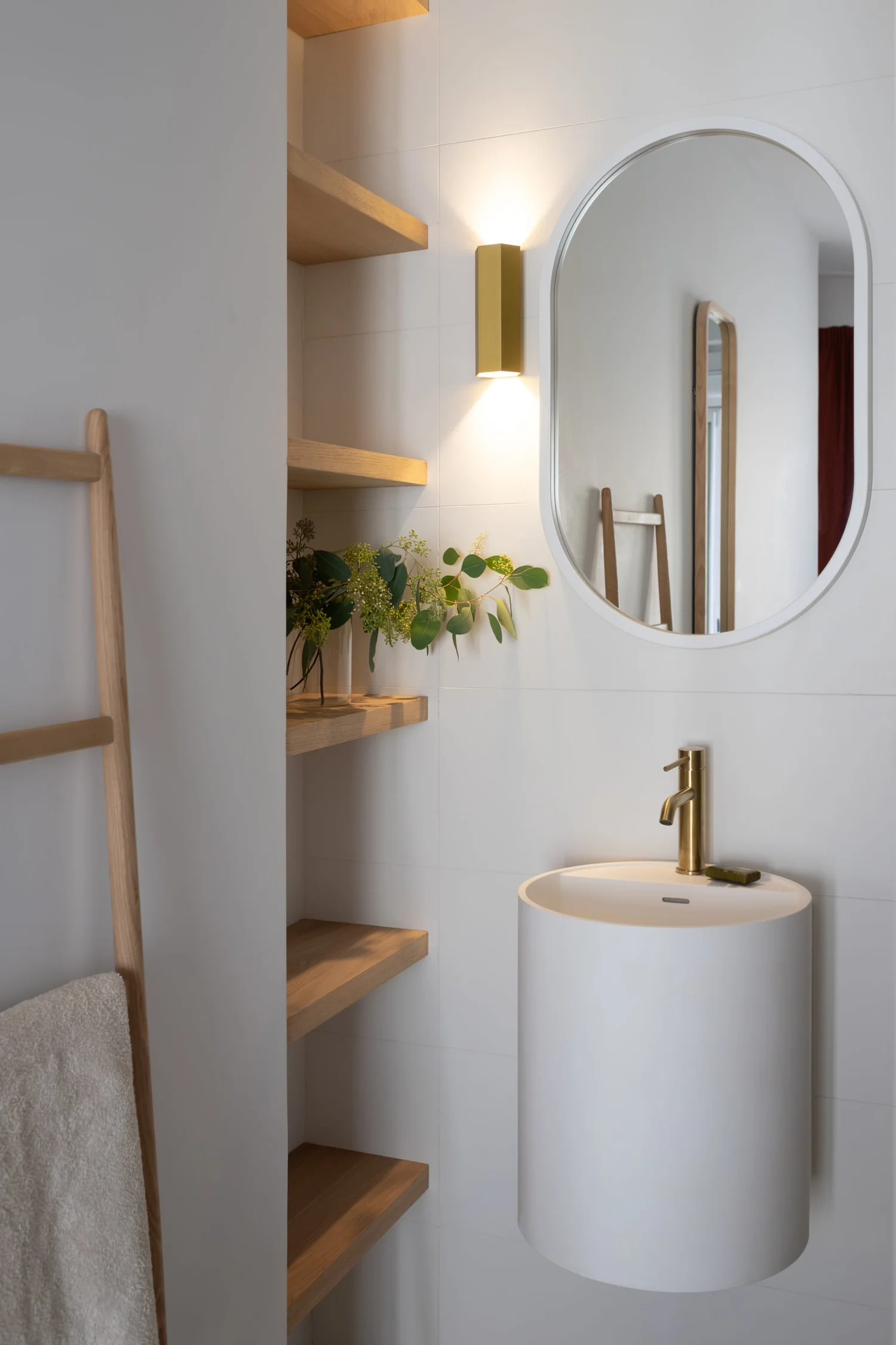 Salle de bain avec miroir ovale, lavabo blanc mural, robinet doré, étagères en bois, plante verte, luminaire mural doré