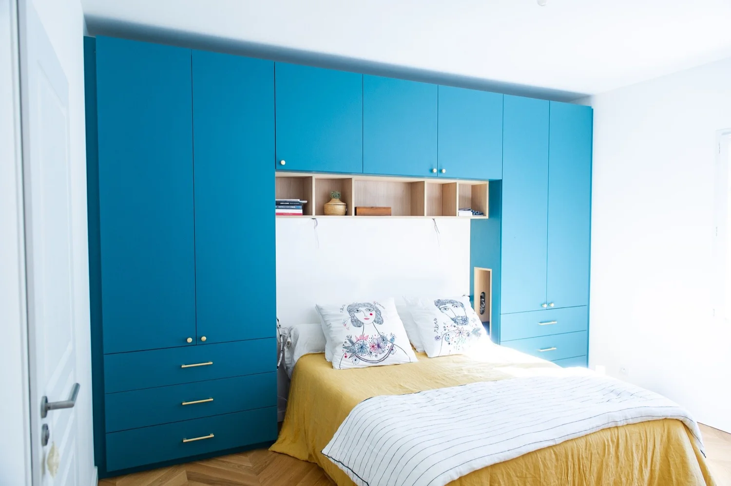 Chambre avec tête de lit intégrée, armoire bleue encadrant un espace de rangement, drap jaune, oreillers avec dessins de femmes, étagères avec livres et objets décoratifs.