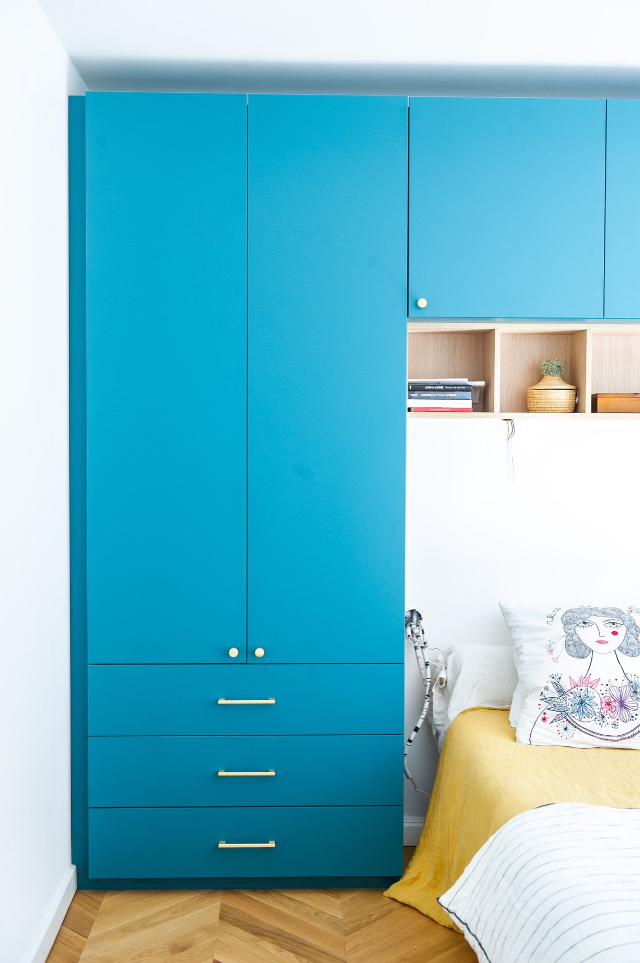 Armoire bleue dans une chambre, partiellement visible, avec un lit à côté ayant une couette jaune et un coussin décoratif blanc avec dessin.