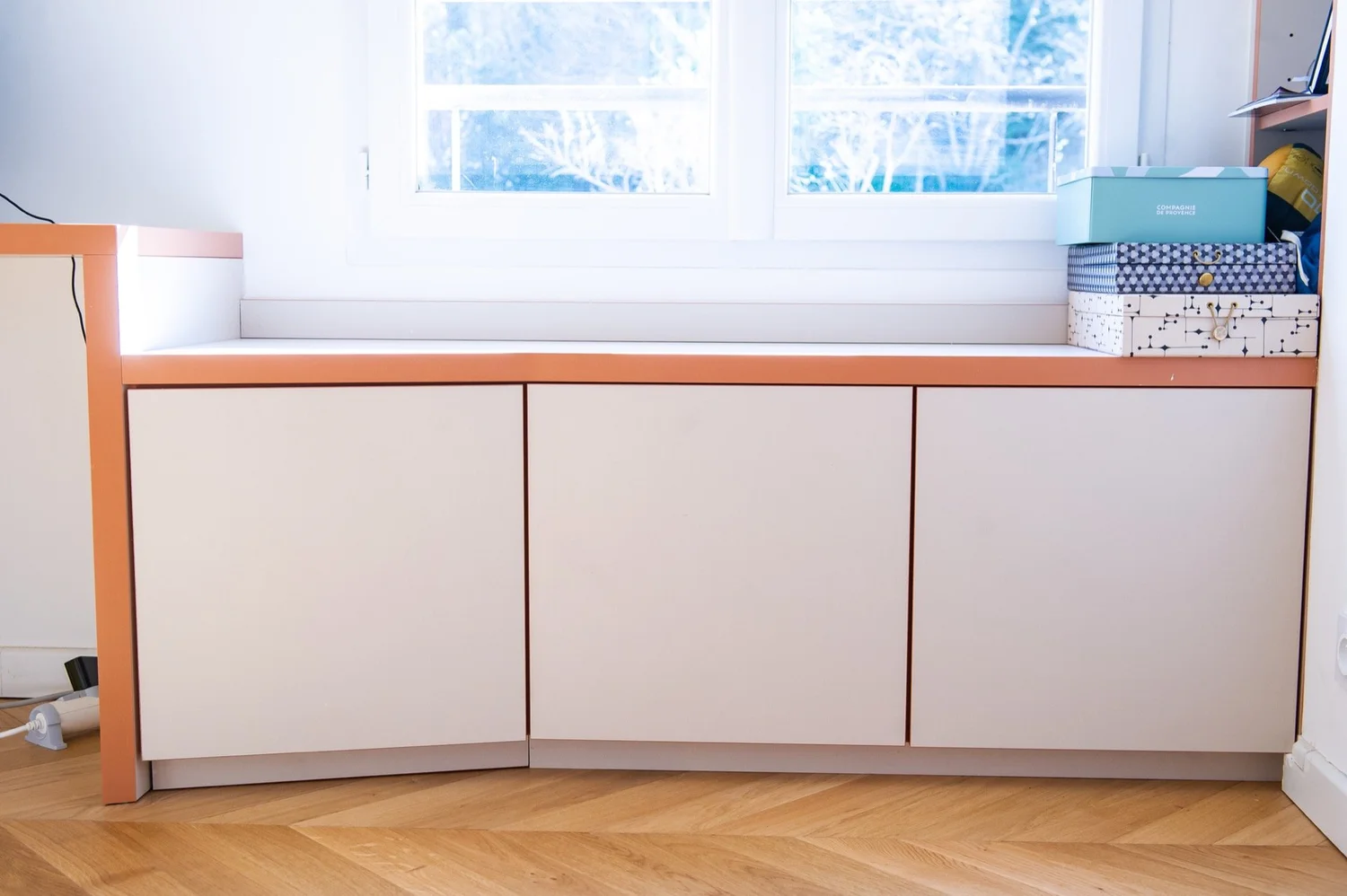 Buffet blanc avec toiture rose clair sous une fenêtre, sur un parquet en bois, avec des boîtes de rangement sur le côté droit.