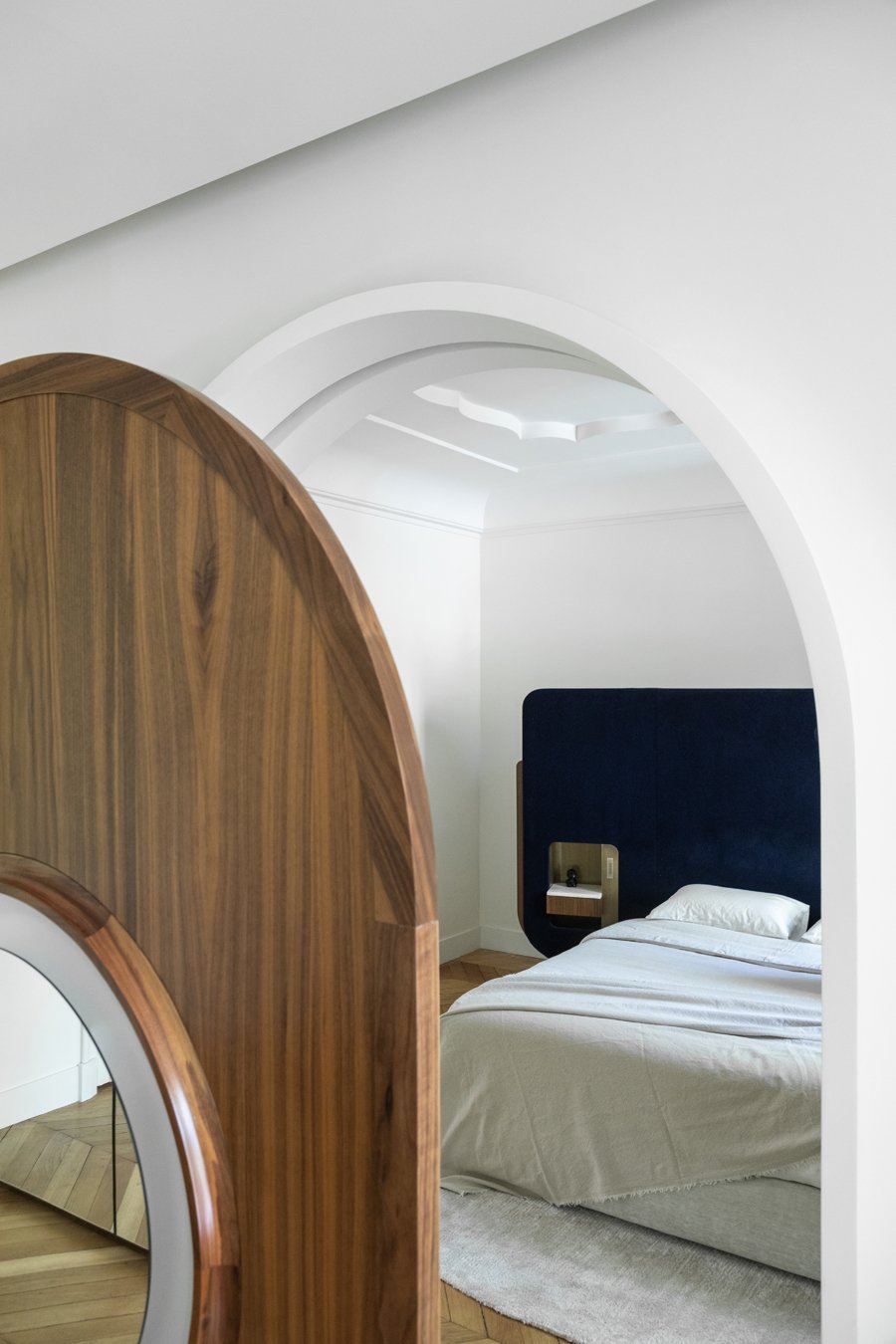 Chambre moderne avec lit à tête bleue, placard en bois et mur blanc, vue par une arche architecturale.