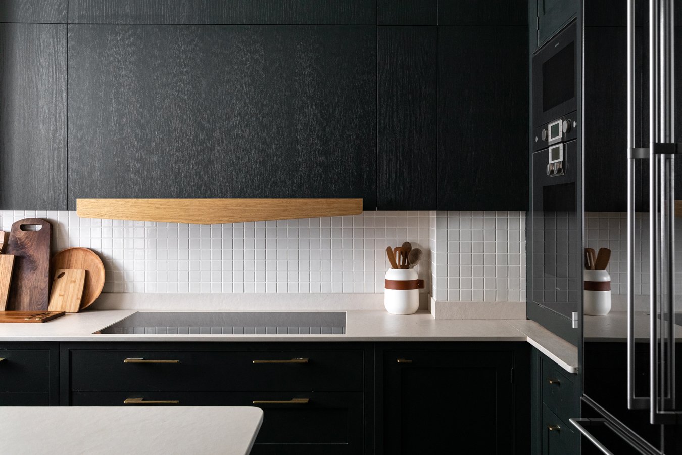 Côté d'une cuisine moderne avec des plans de travail en quartz, des armoires noires avec des poignées dorées, un mur de carrelage blanc et des planches de bois. Un vase blanc avec des ustensiles de cuisine en bois se trouve sur le plan de travail.