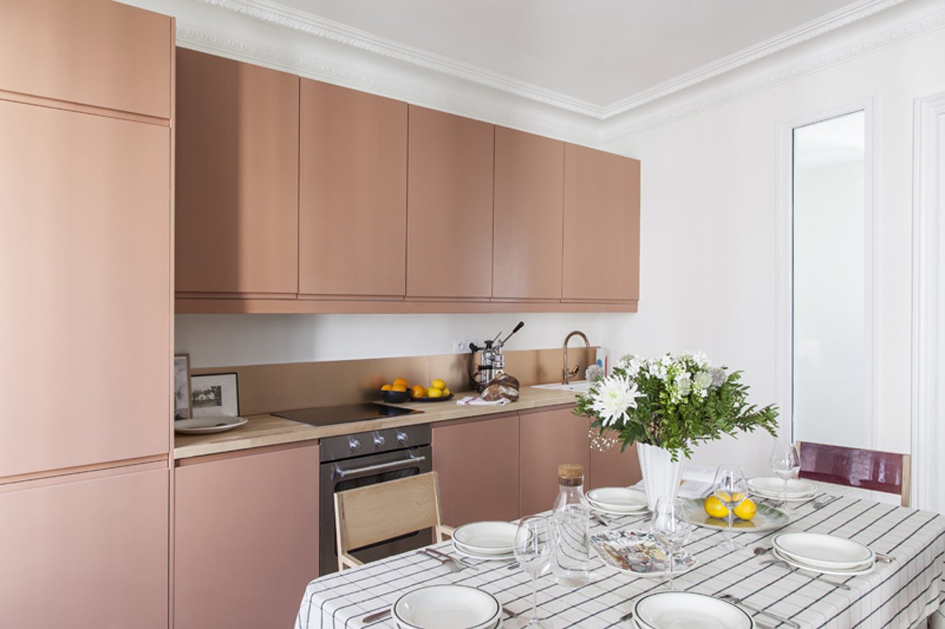 Cuisine moderne avec armoires roses, table de salle à manger avec vaisselle, fleurs dans un vase, et fruits.