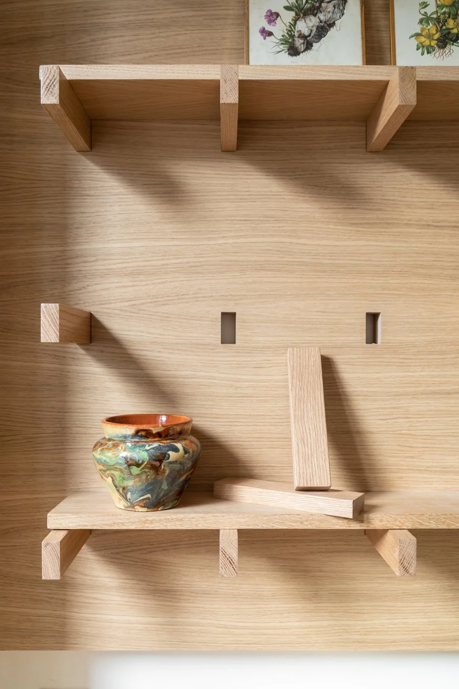 Une étagère en bois avec un pot en céramique coloré et deux blocs de bois, avec des cadres de fleurs au-dessus.