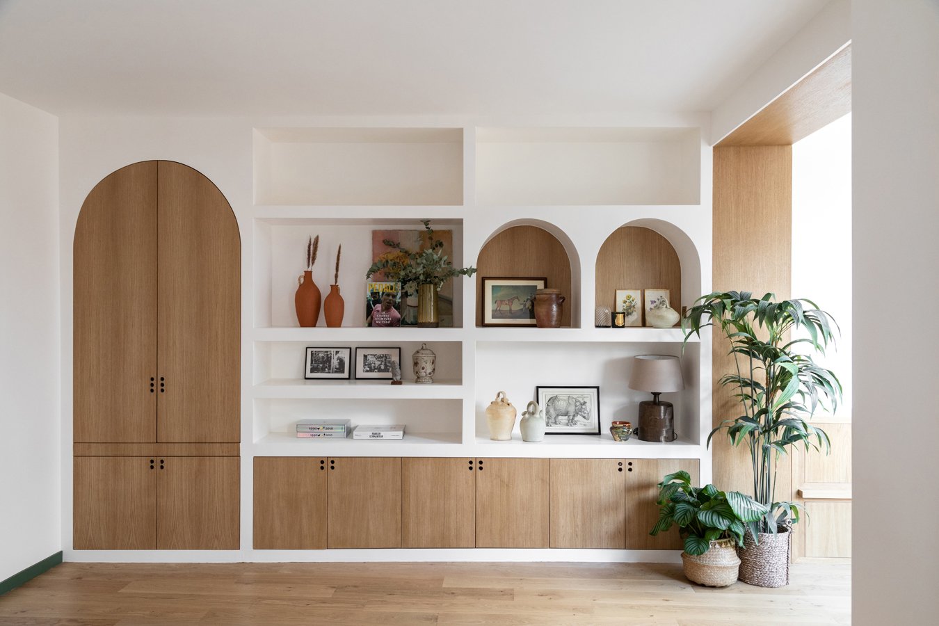 Une étagère en bois intégrée dans un mur blanc, décorée avec des plantes, des cadres, des vases, et des objets décoratifs, avec un grand potted plant à droite et un sol en bois.