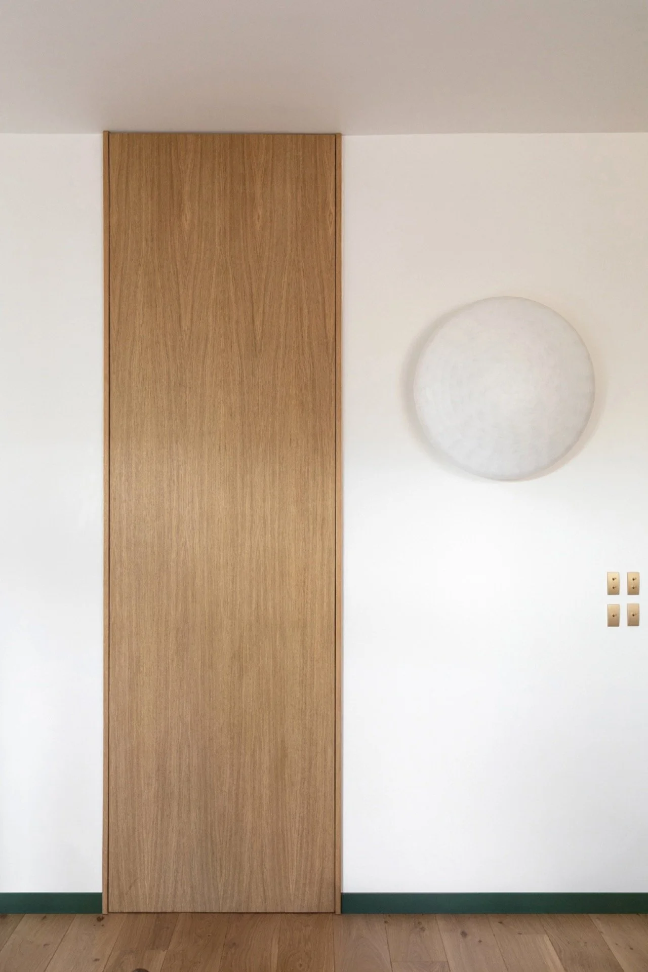Portail en bois intégré dans un mur blanc avec une décoration circulaire en relief à droite et quatre prises électriques en dessous.