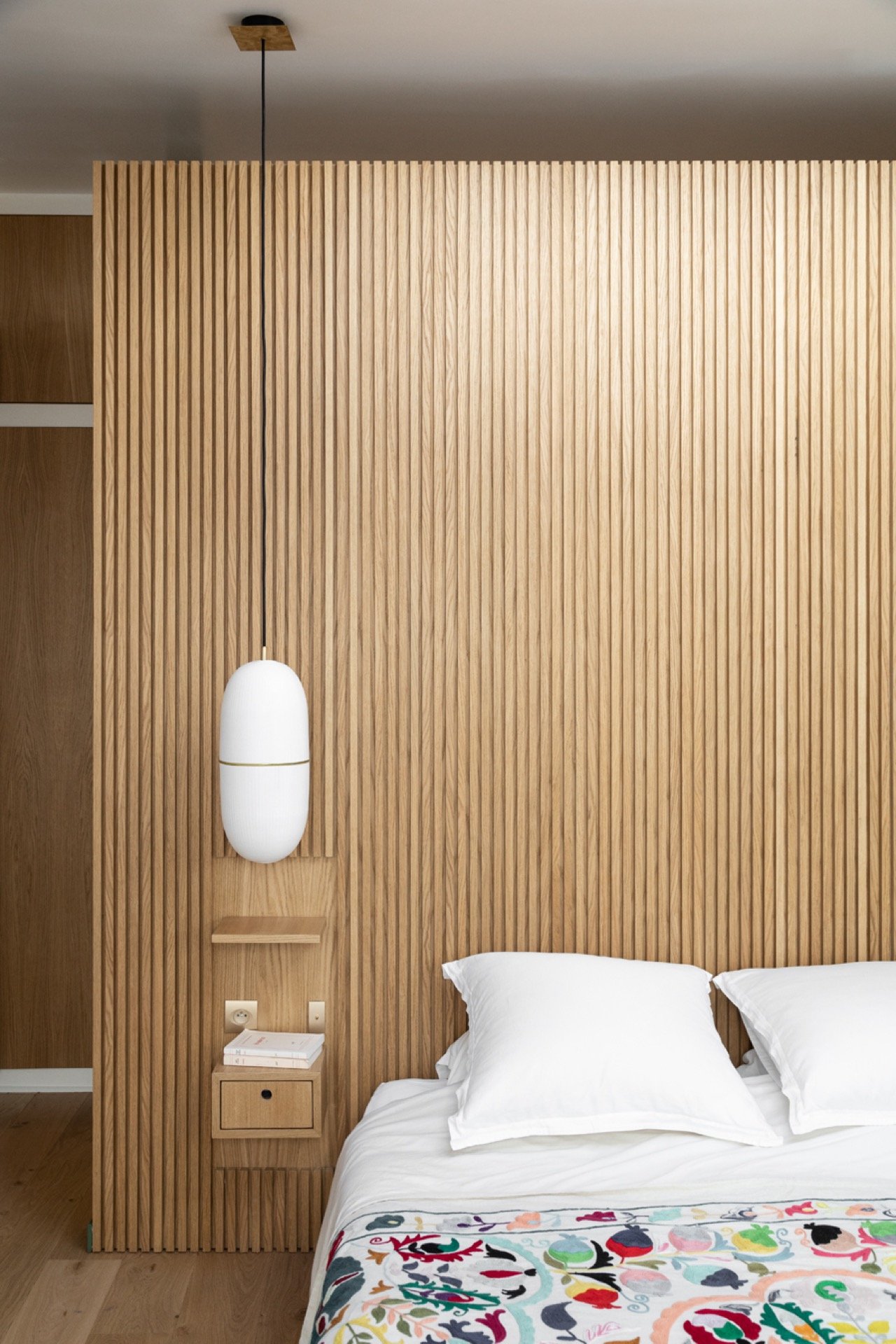 Mur en bois clair avec tête de lit en bois, lampe suspendue blanche, petite étagère avec livres, chêne clair, lit avec draps blancs et couverture colorée que à moitié visible.