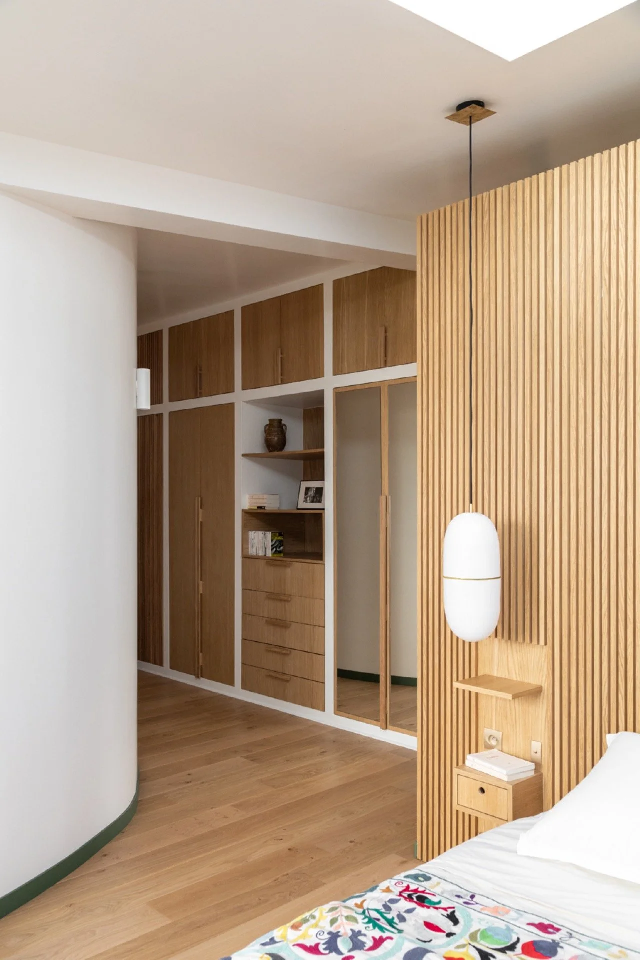 Intérieur d'une chambre avec lit en bois, une tête de lit en bois à rayures, suspension blanche au-dessus d'une petite table avec livres, et un mur avec des placards en bois et étagères