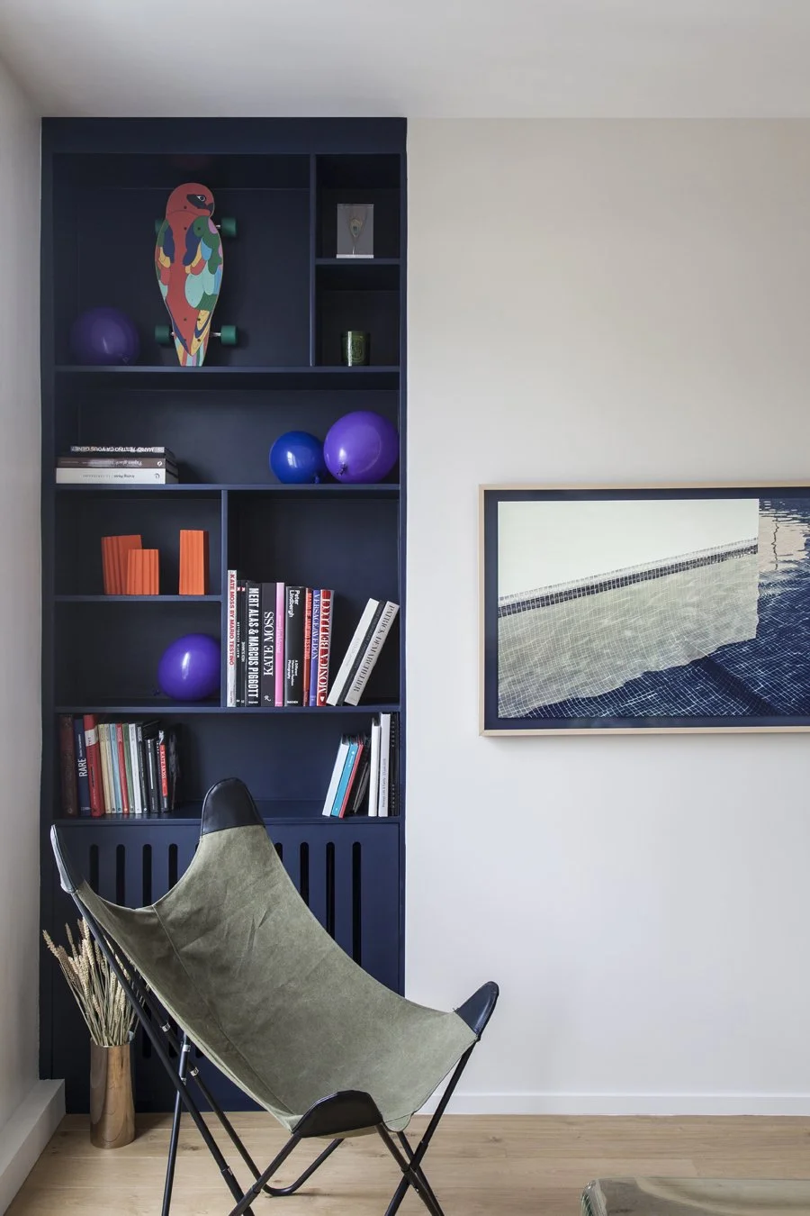 Un coin salon avec une étagère noire pleine de livres, ballons et décoration, un fauteuil pliant vert avec un repose-pied, un vase avec des herbes sèches, une grande peinture abstraite sur le mur.