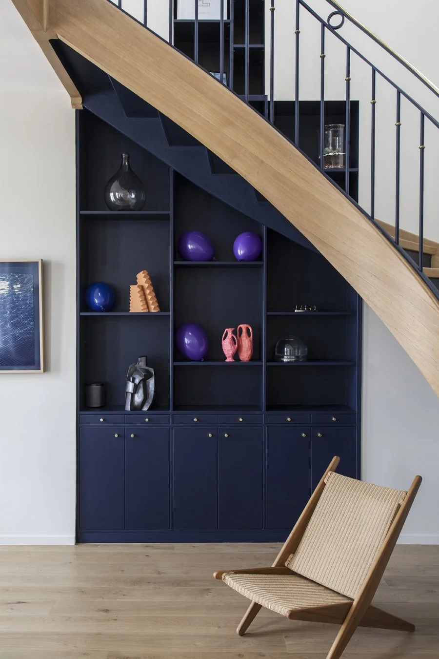 Étagère intégrée sous un escalier avec objets décoratifs colorés, chaise en bois avec assise en tissu, et partie de l'escalier en bois et métal.