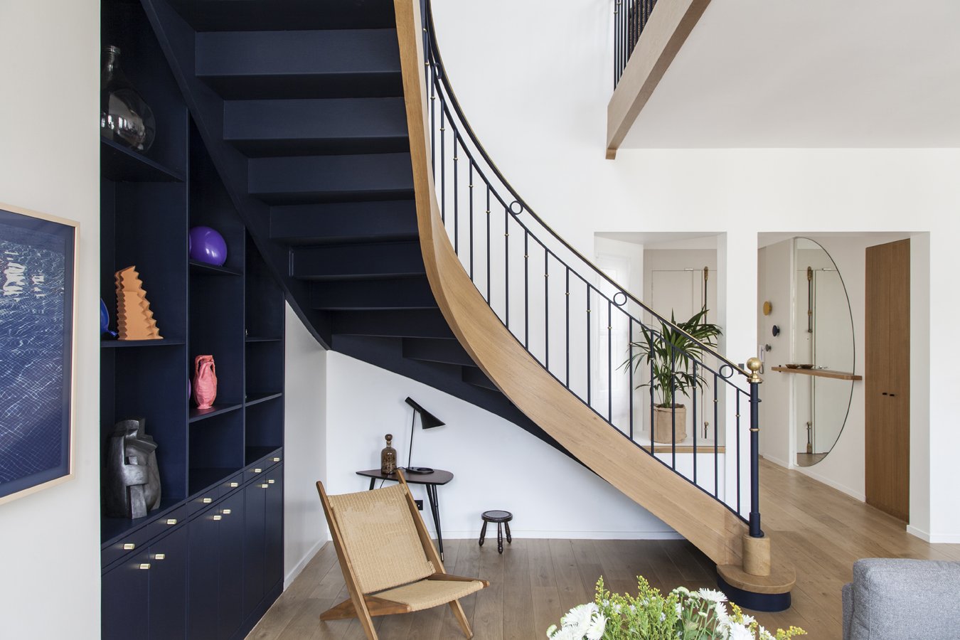 Intérieur moderne avec escalier en bois, garde-corps en métal, étagères bleues avec objets décoratifs, fauteuil en bois, plante verte, miroir mural, parquet en bois clair.
