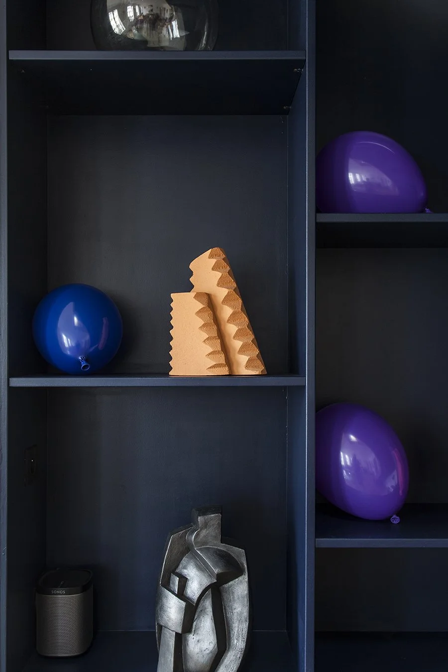 Étagère avec un ballon bleu, deux éponges en forme de pyramide orange, une sculpture en métal argenté, une enceinte noire, et deux ballons violets sur les étagères.