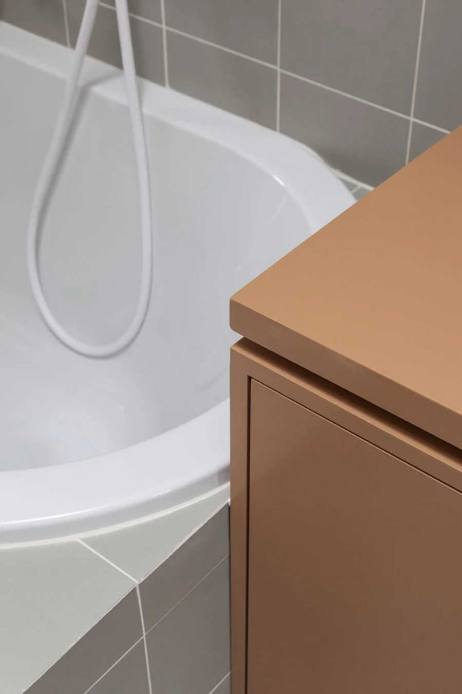 Baignoire blanche avec un robinet de douche et un mur en carrelage gris, partiellement visible d'une armoire ou meuble en bois de couleur beige