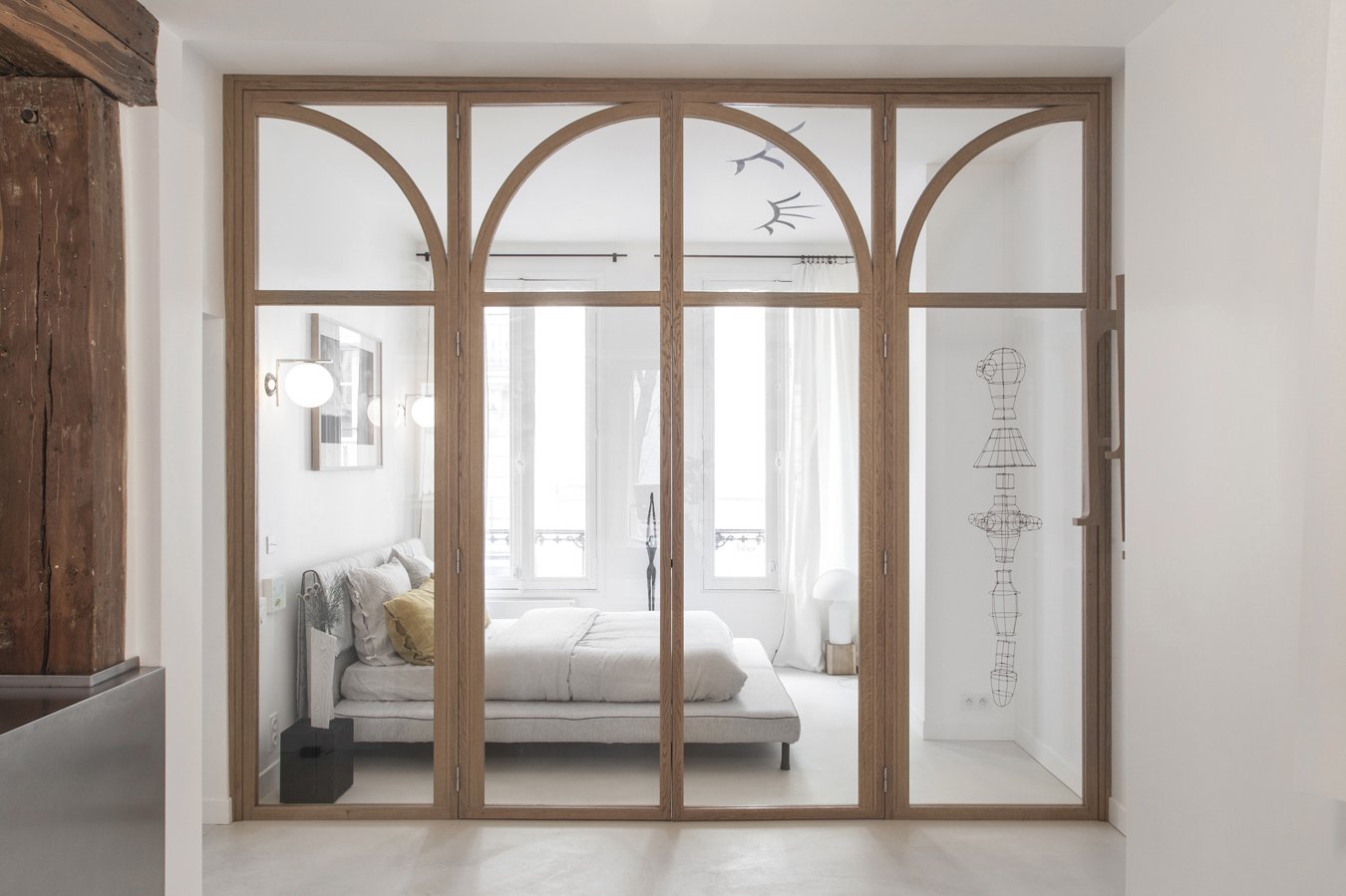 Une chambre moderne visible à travers une cloison en bois avec vitres, avec un lit blanc, des fenêtres grandes laissant entrer beaucoup de lumière, et un dessin en fil de fer d'une tête et d'un corps humanoïdes sur le mur à droite.