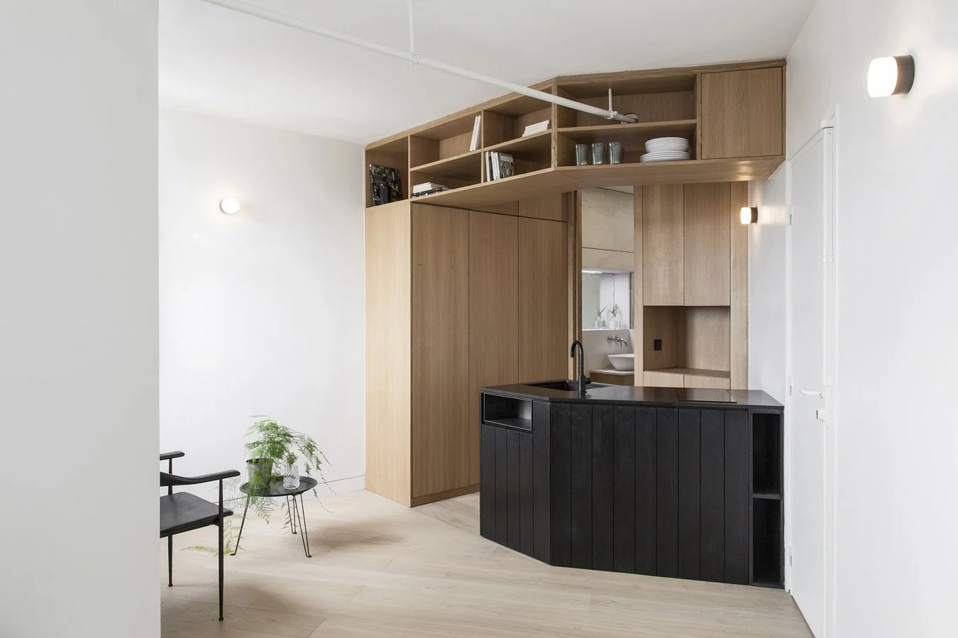 Intérieur d'une cuisine moderne avec unité en bois clair, comptoir noir, étagères ouvertes en bois, petite table noire avec une plante, chaise noire, murs blancs