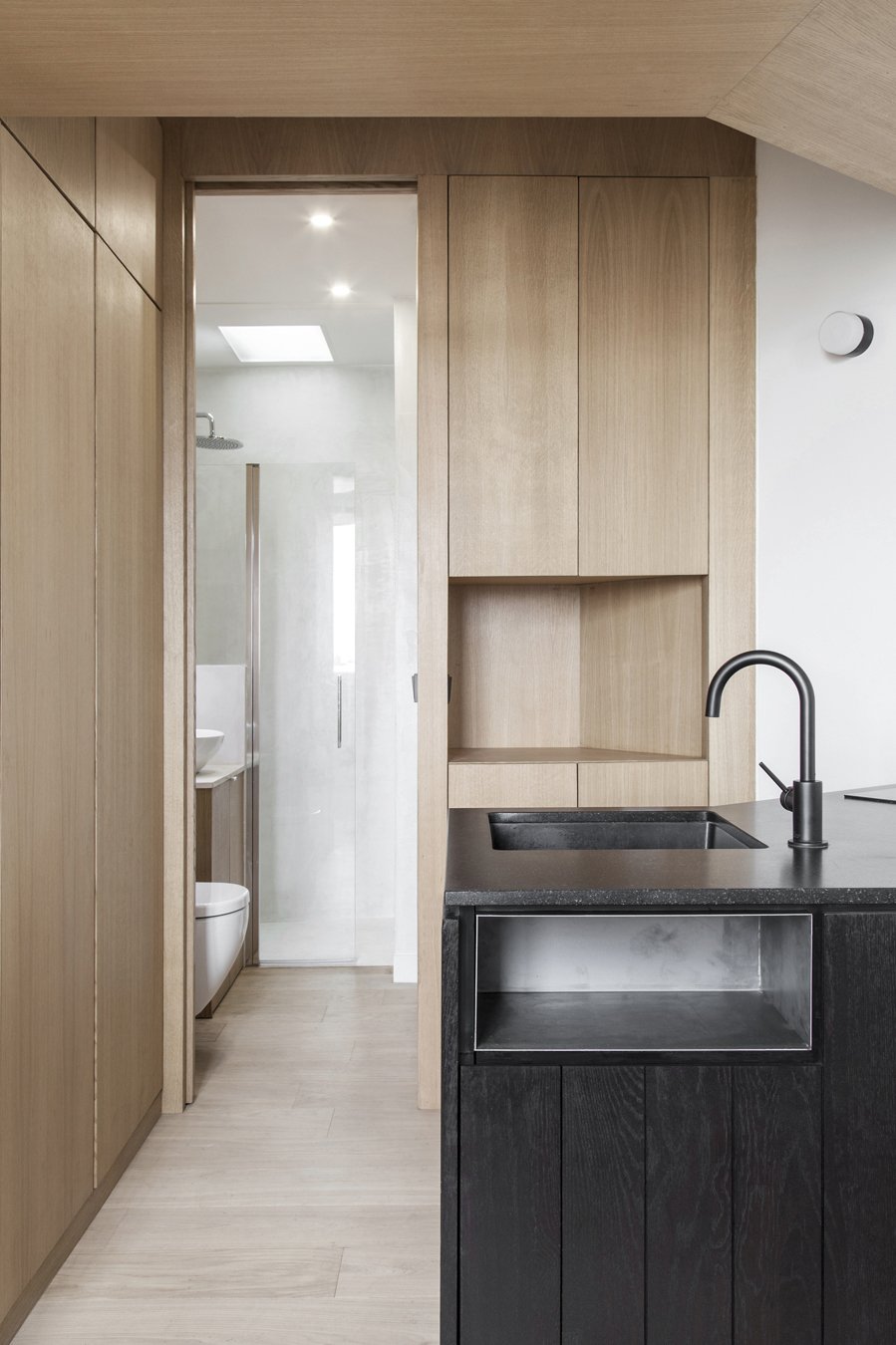 Cuisine moderne avec plan de travail noir, évier noir et mobilier en bois clair. En arrière-plan, salle de bain avec douche et toilettes visibles.