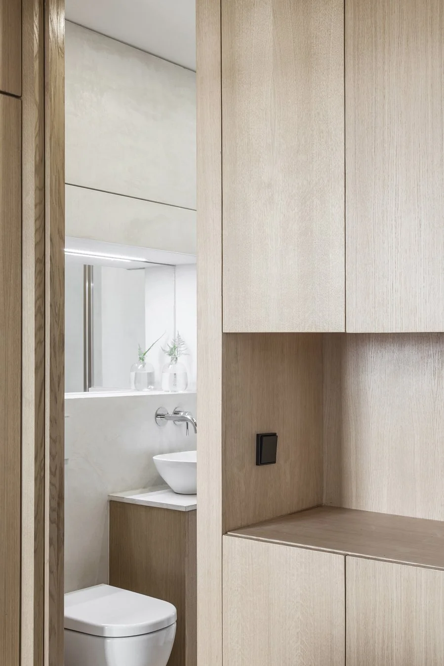 Vue partielle d'une salle de bain moderne avec mobilier en bois clair, lavabo blanc, miroir, et décorations minimalistes avec des vases et des plantes.