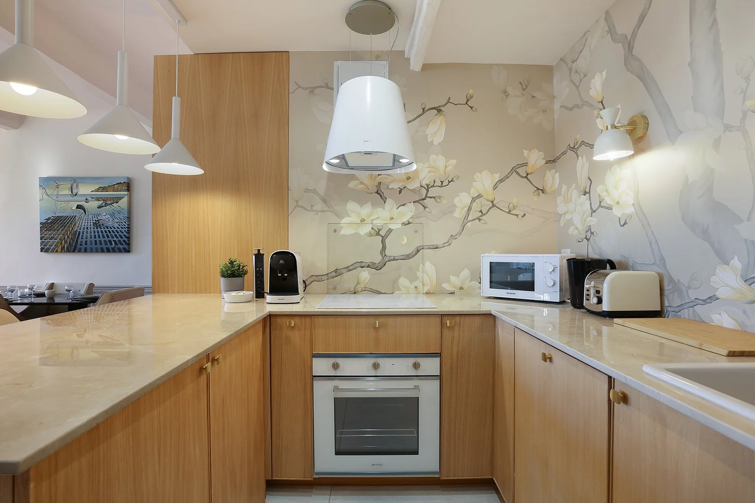 Cuisine moderne avec plans de travail en marbre, mur décoré avec une fresque de branches de magnolia, appareils électroménagers blanc et noir, et éclairage suspendu blanc.