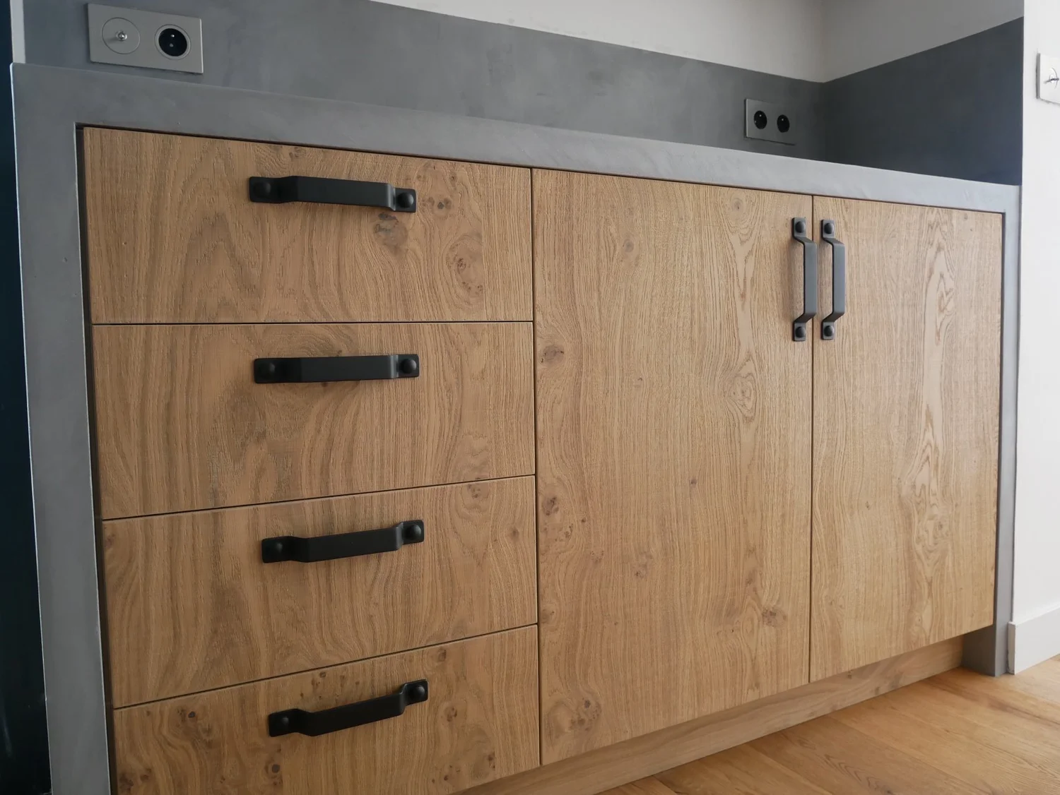 Meuble en bois clair avec cinq tiroirs à gauche et deux portes à droite, poignées noires, sur un comptoir gris avec deux prises électriques et un câble visible.