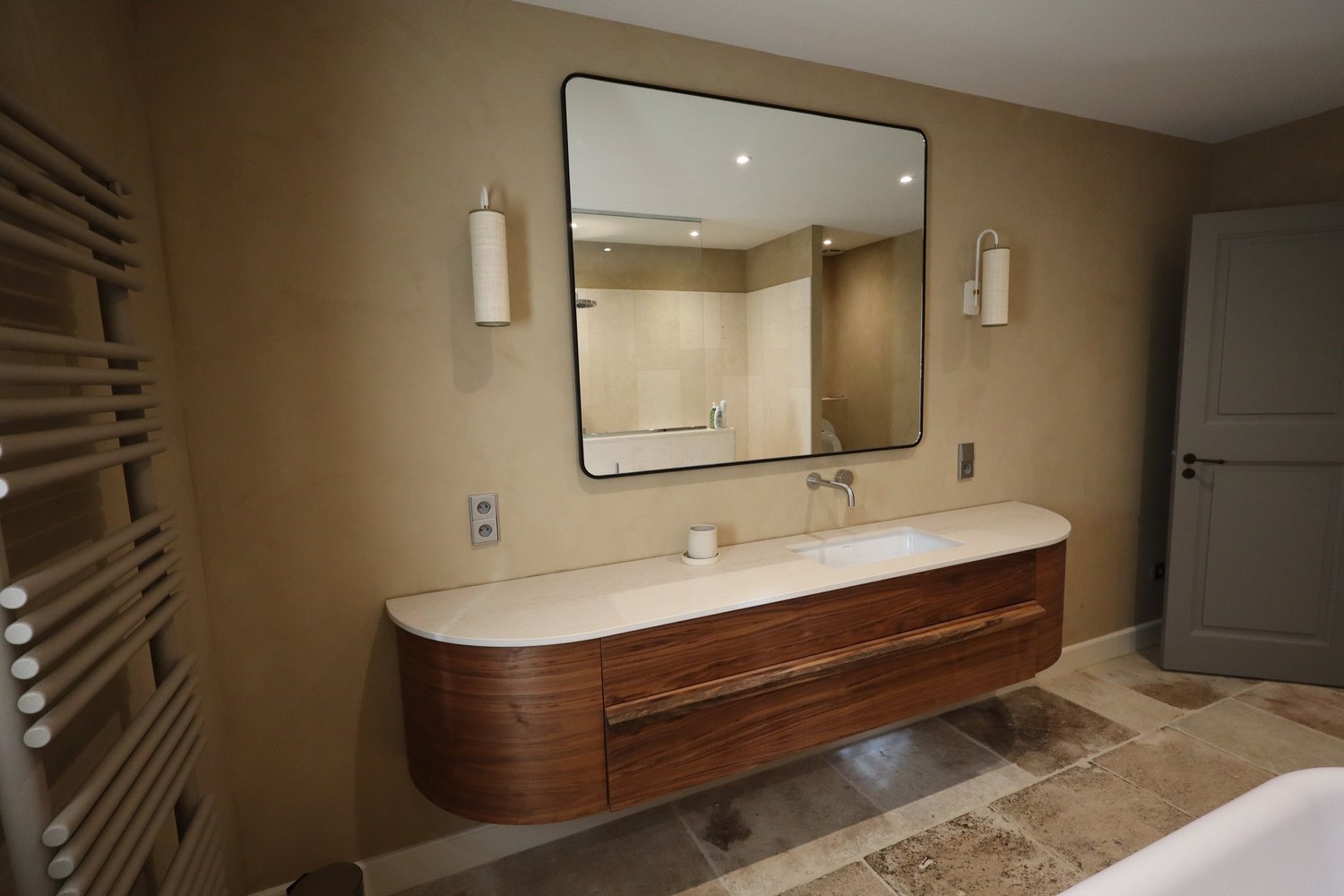 Un miroir carré au-dessus d'une vasque intégrée à un meuble en bois avec un comptoir en pierre, dans une salle de bain avec des murs beige et un sol en carrelage. Deux appliques murales de chaque côté du miroir. Le reflet montre une douche et un WC.