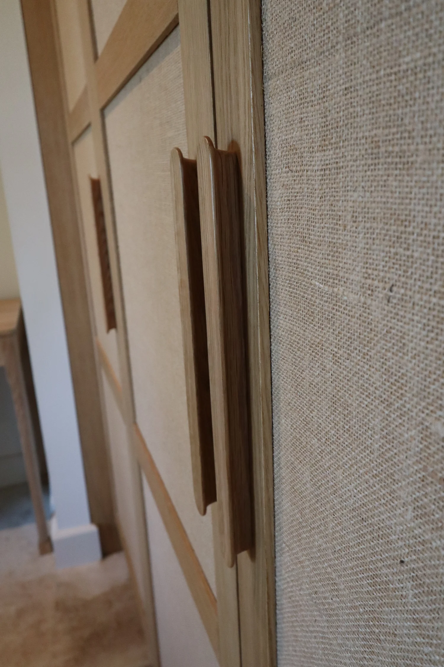 Une image montrant un meuble en bois avec deux poignées en bois, fixé à un mur recouvert de tissu beige.