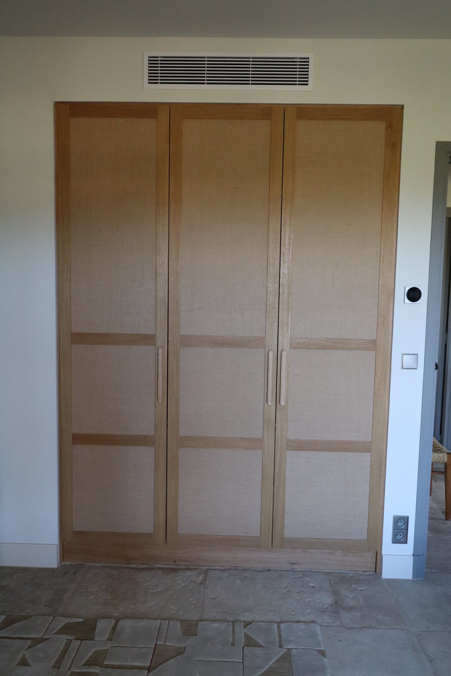 Une armoire en bois avec des portes coulissantes, située à l’intérieur d’une pièce, avec un plafond à ventilation et des prises électriques sur le mur adjacent.