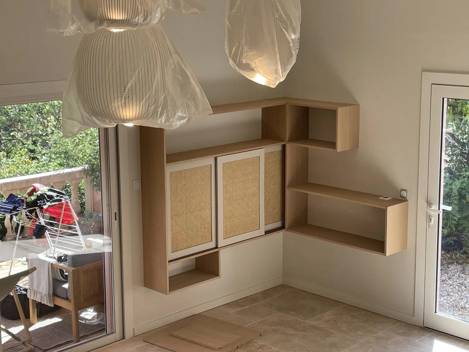 Partie intérieure d'une pièce avec des étagères en bois, une porte-fenêtre donnant sur un balcon avec du linge suspendu, et un luminaire avec des lampes en plastique.