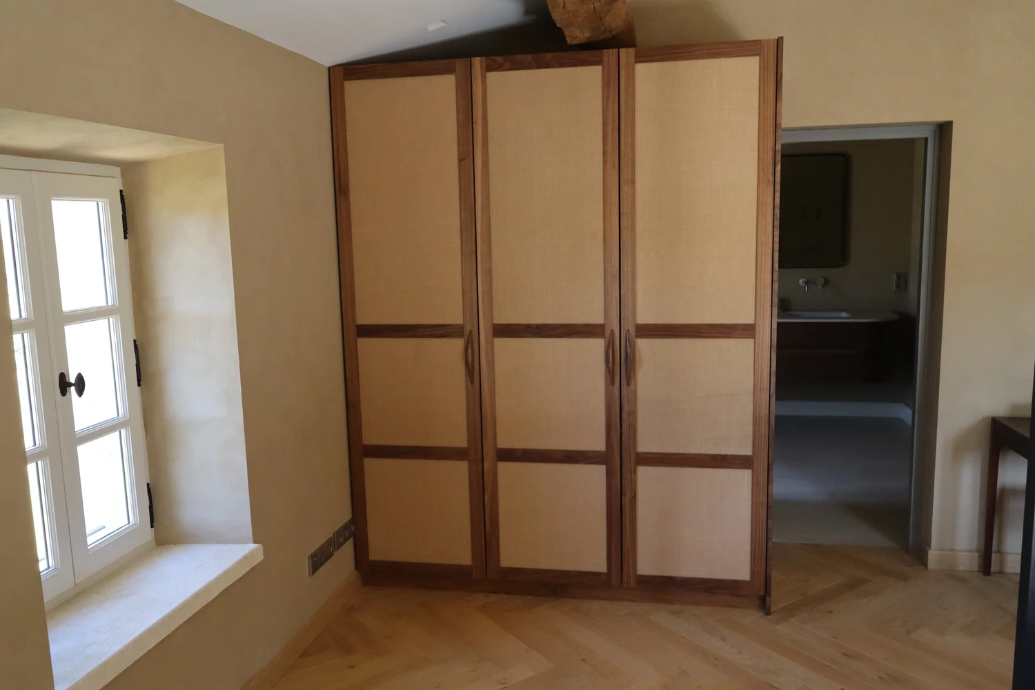 Une pièce avec un coffre en bois et toile beige, une fenêtre avec volets blancs, et une porte menant à une salle de bain.