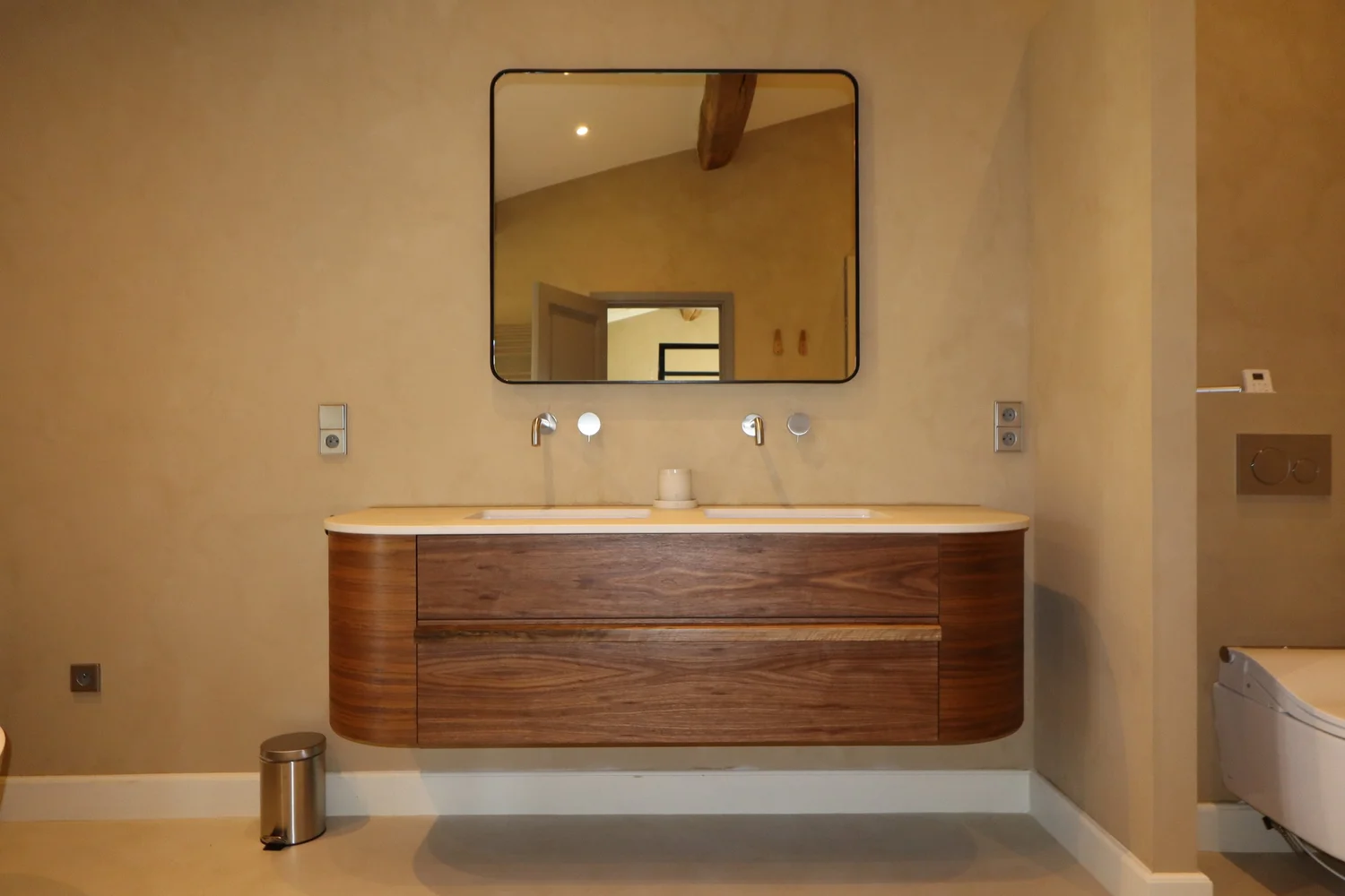 Salle de bain avec un meuble double lavabo en bois, un miroir rectangulaire, une paroi de douche en verre partielle et des murs beige, avec un petit poubelle en acier et une toilette à droite.