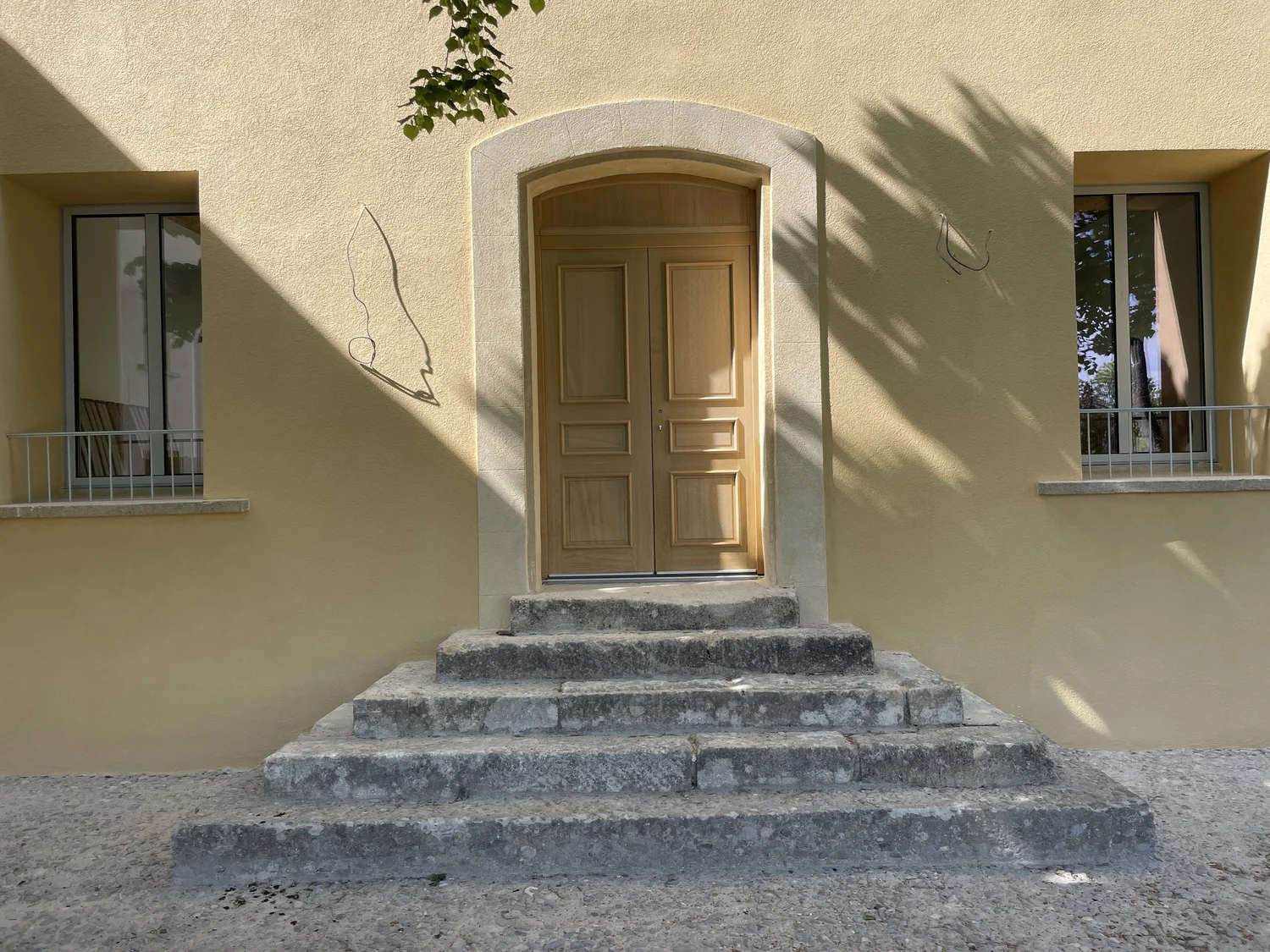 Façade d'une maison avec une porte en bois entourée de murs jaunes, deux fenêtres avec volets, et des escaliers en pierre menant à la porte, avec des ombres projetées par des arbres.