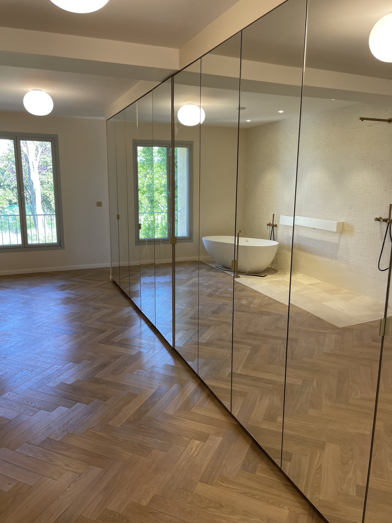 Salle de bain moderne avec baignoire autoportante et murs en mosaïque, séparée par un mur de miroirs du reste de la pièce, avec fenêtres laissant entrer beaucoup de lumière naturelle.