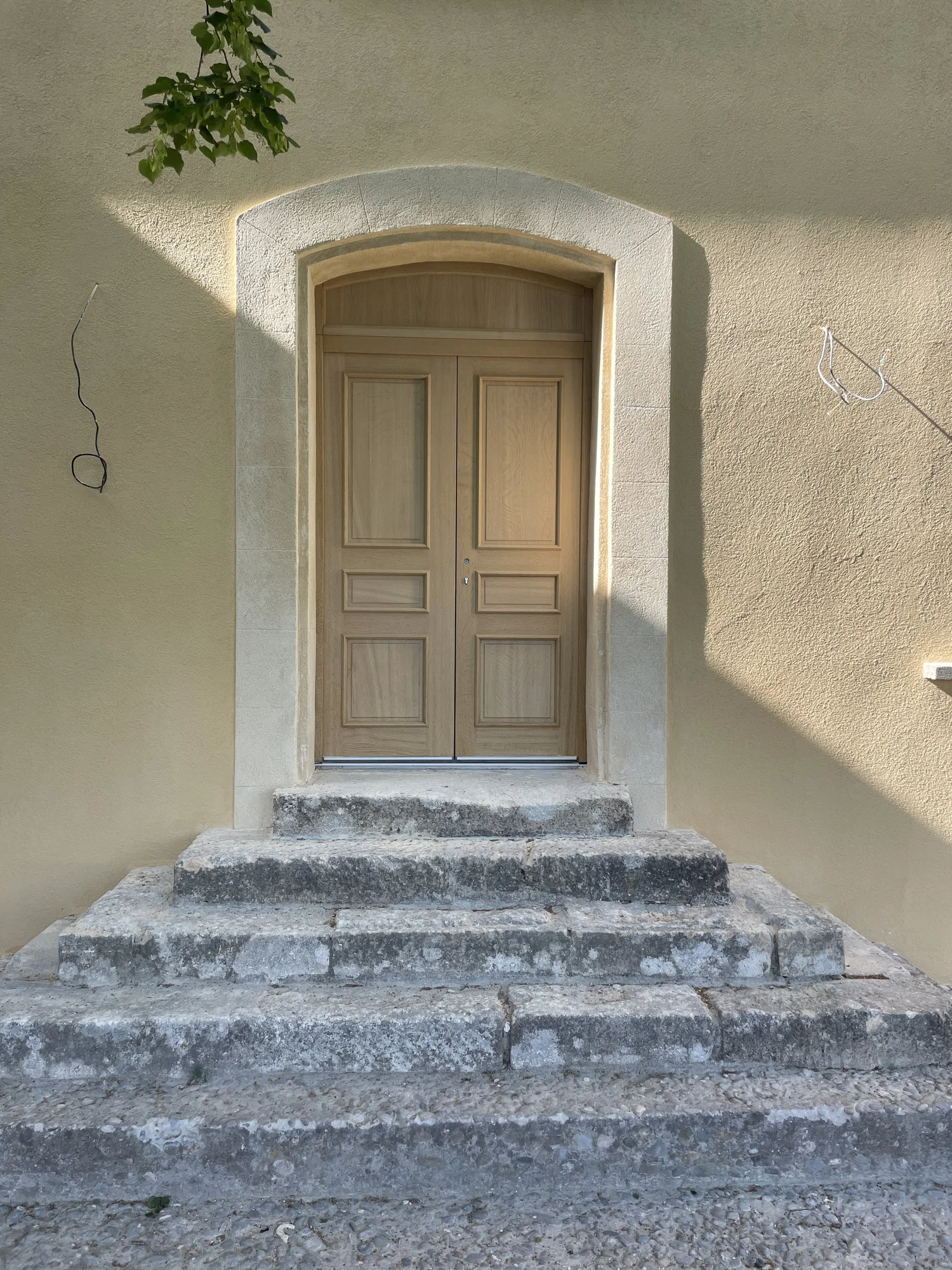 Porte en bois naturel avec étage en pierre, accessible par trois marches en pierre, sur un mur beige clair, avec une branche d'arbre en haut à gauche.