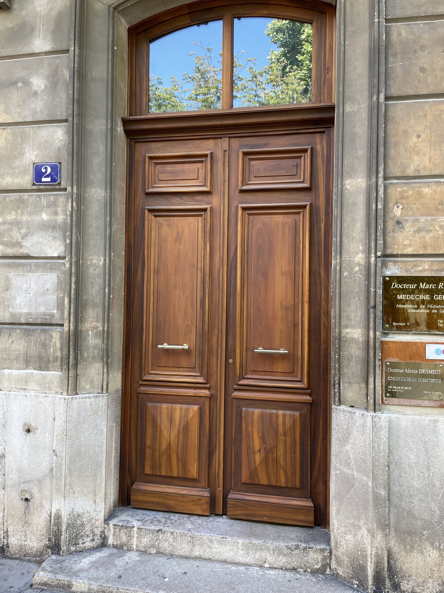Porte en bois avec deux poignées en métal, située dans une façade en pierre avec une plaque bleue portant le numéro 2.
